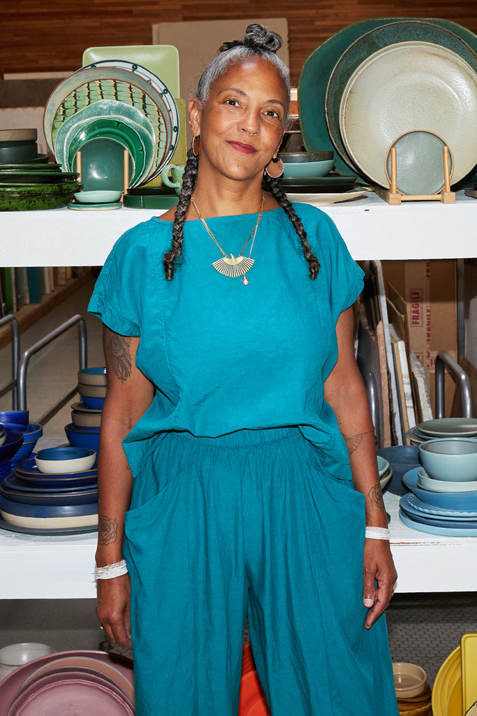 Model wears Jones Top in Ocean and Larkin Pant in Ocean, standing in front of shelves holding colorful bowls and plates.