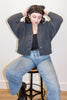 Person wearing a dark gray Lino textured cardigan and blue jeans sitting on a stool.