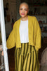 Model wearing Lino Cardigan in Chartreuse, Minnie Tank in White, and Jessie Stripe Skirt in Dijon, standing in foreground of living room space