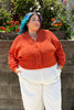 Person wearing an orange Lino sweater and white Cadence pants standing against a stone wall with flowers in the background
