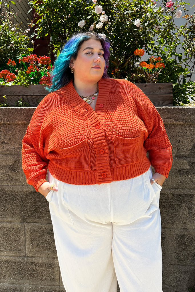 Person wearing an orange Lino sweater and white Cadence pants standing against a stone wall with flowers in the background