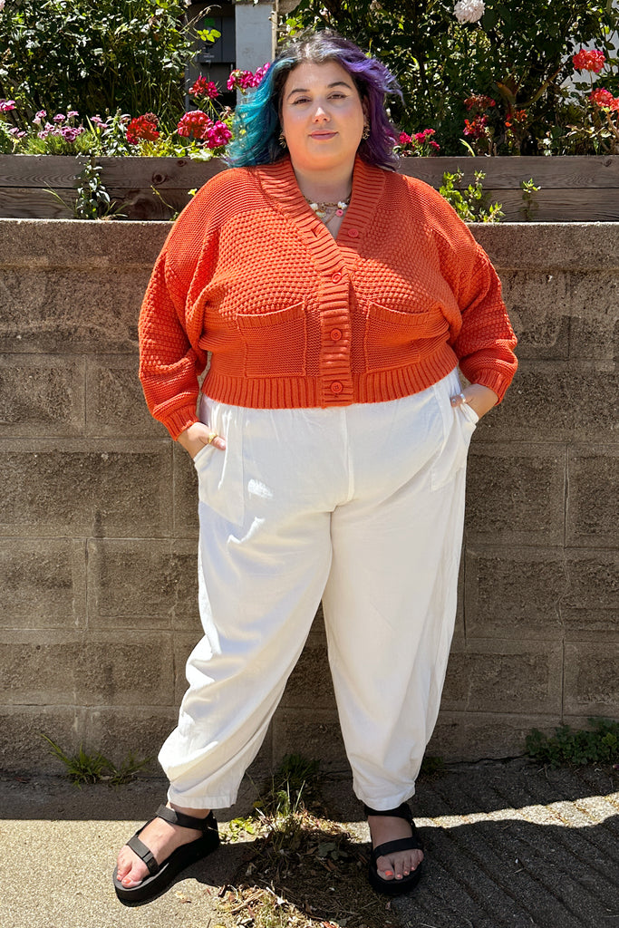 Person wearing an orange Lino sweater and white Cadence pants standing against a stone wall with flowers in the background