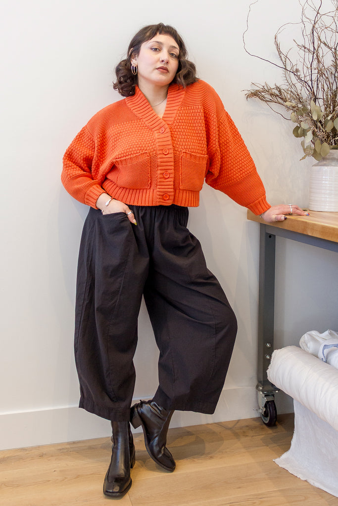 Model wearing Lino Cardigan in Tomato and Larkin Pant in Black, leaning against a table in front of white background