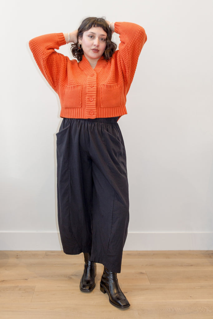 Model wearing Lino Cardigan in Tomato and Larkin Pant in Black, standing with arms raised above head, in front of white background