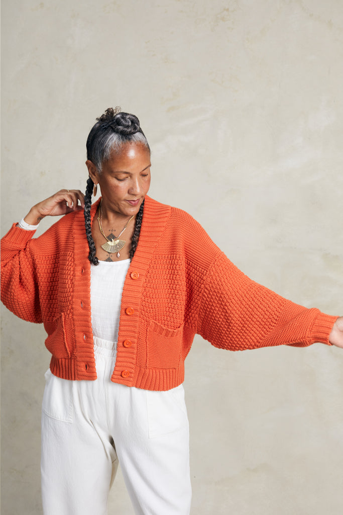 Lino Cardigan in Tomato and Cadence Pant in White on model with arm extended out to the side, in front of white background