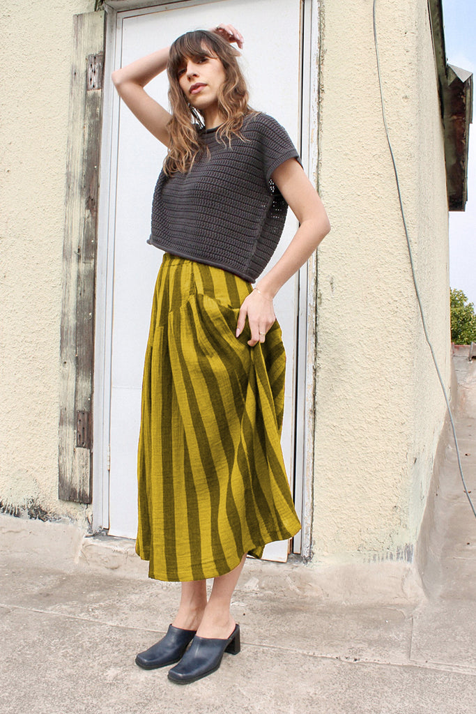 Model wearing Niko Sweater in Charcoal and Jessie Stripe Skirt in Dijon in front of white door of light yellow building