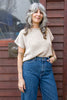 Model wearing Niko Sweater in Oat and blue jeans in front of wooden building