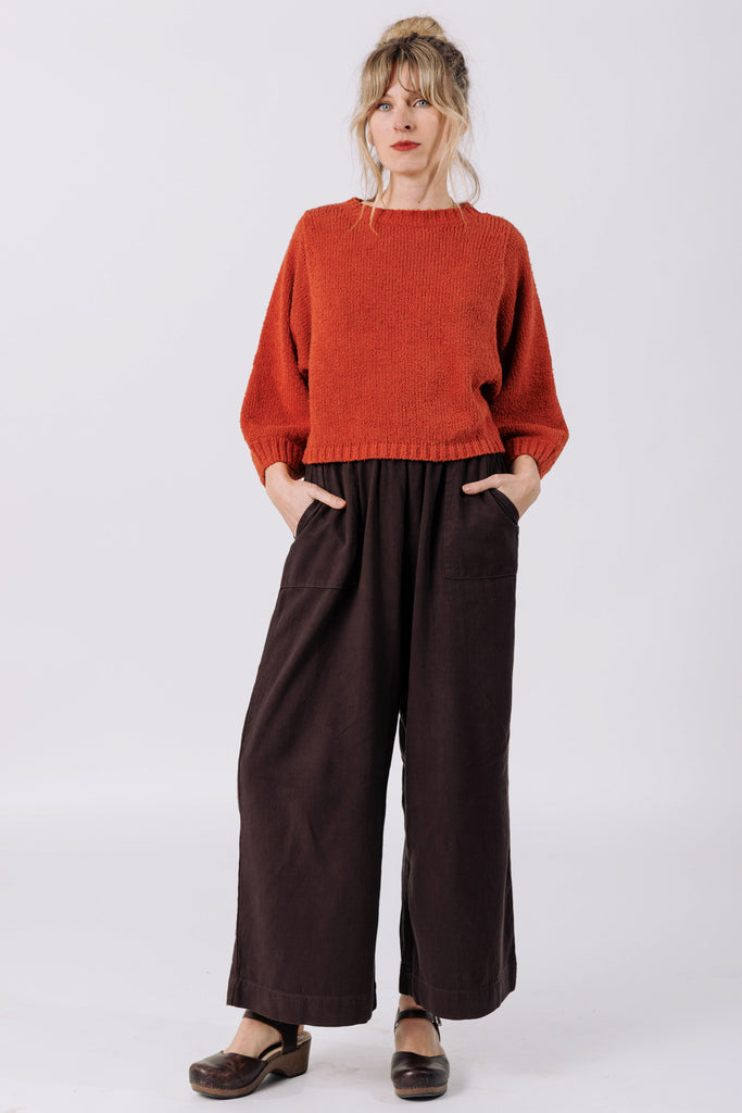 Front view of model wearing Nova Sweater in Paprika and Atlas Pant in Chocolate, standing  in front of white wall