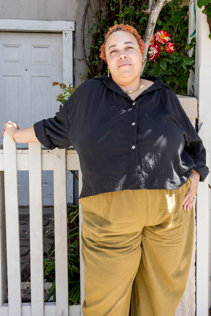 Model wearing Cropped Poet Blouse in Black and Cadence Pants in Dijon, leaning against white fence