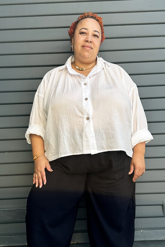 Model wearing Cropped Poet Blouse in White and Cadence Pant in Black, in front of dark green paneled wall