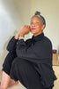 Side view of model wearing Cropped Poet Blouse in Black, sitting in front of white walls