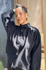 Model wearing Cropped Poet Blouse in Black, with arm raised to head, in front of grey and wooden building 