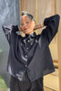 Model wearing Cropped Poet Blouse in Black, with arms raised to head, in front of grey and wooden building 
