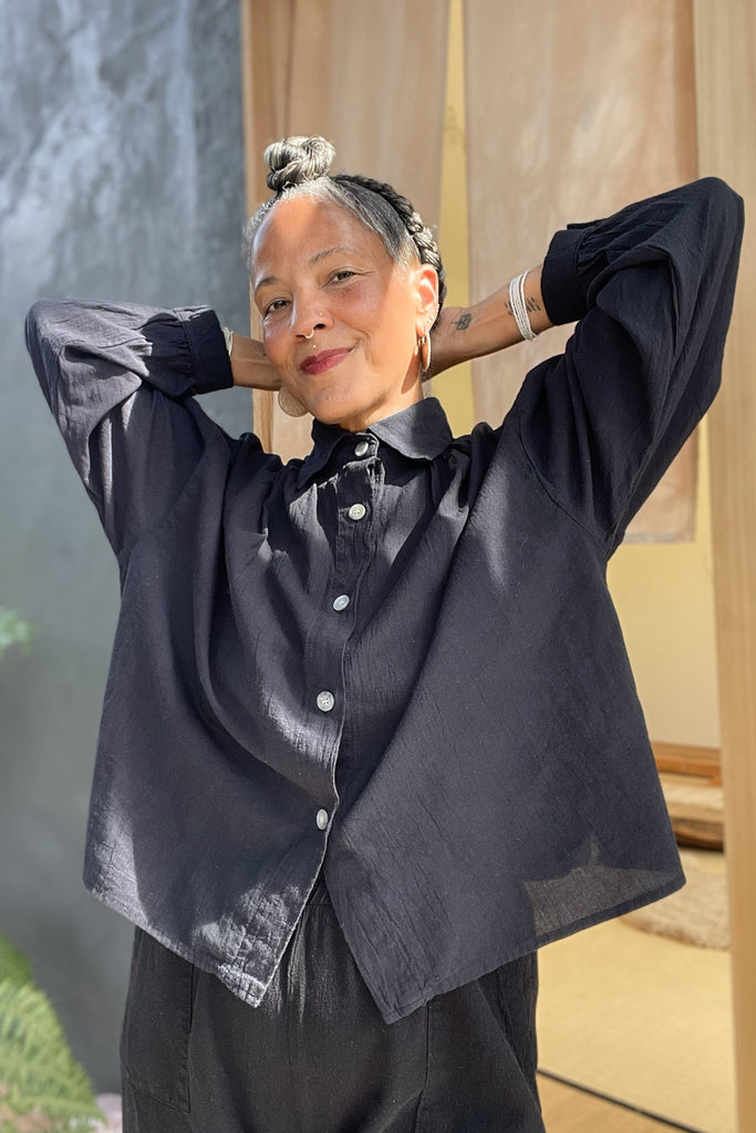 Model wearing Cropped Poet Blouse in Black, with arms raised to head, in front of grey and wooden building 