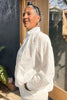 Side view of model wearing Cropped Poet Blouse in White and Cadence Pant in White, standing with hand in pocket in front of grey and wooden building