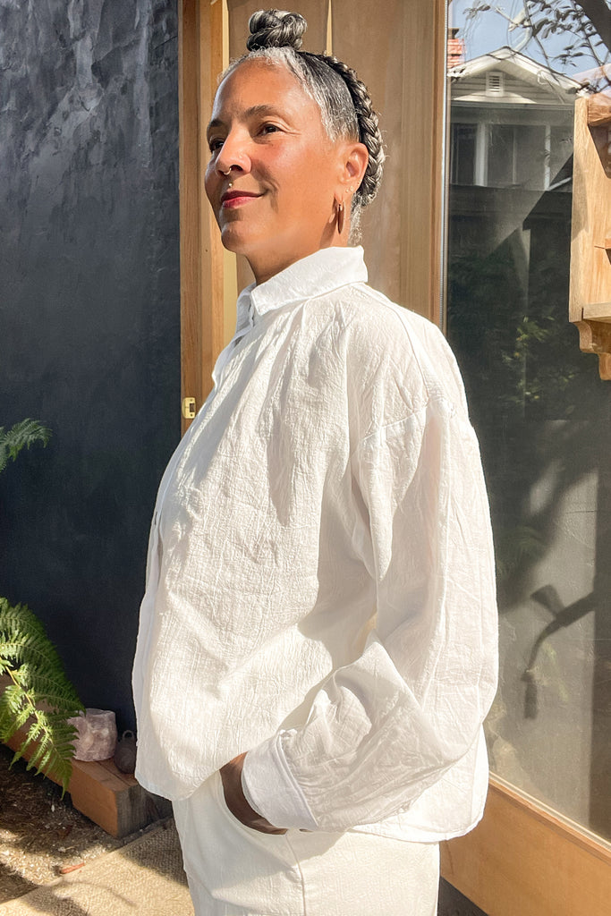 Side view of model wearing Cropped Poet Blouse in White and Cadence Pant in White, standing with hand in pocket in front of grey and wooden building
