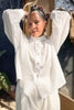 Model wearing Cropped Poet Blouse in White and Cadence Pant in White with hands raised to their head, standing in front of grey and wooden building 