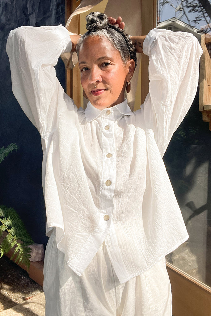 Model wearing Cropped Poet Blouse in White and Cadence Pant in White with hands raised to their head, standing in front of grey and wooden building 