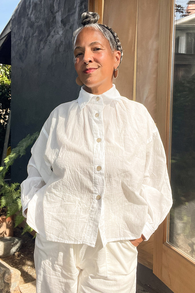 Model wearing Cropped Poet Blouse in White and Cadence Pant in White with hands in pockets, standing in front of grey and wooden building 