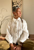 Model wearing Cropped White Poet Blouse in White and Cadence Pant in Fennel, sitting crosslegged in front of small tree and wall art on white wall