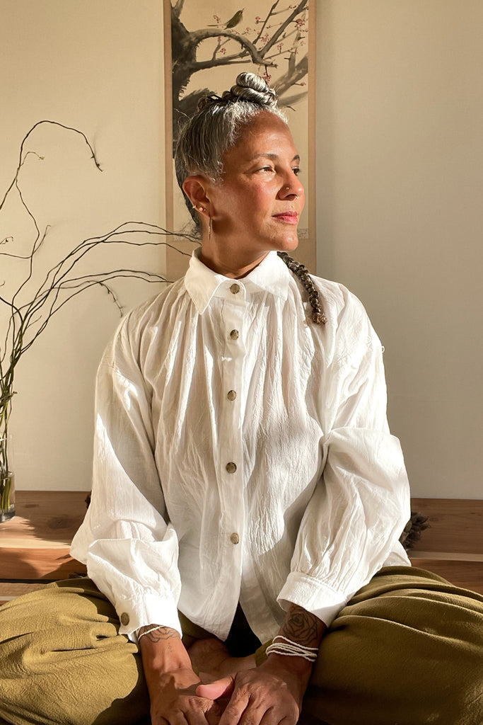 Model wearing Cropped White Poet Blouse in White and Cadence Pant in Fennel, sitting crosslegged in front of small tree and wall art on white wall