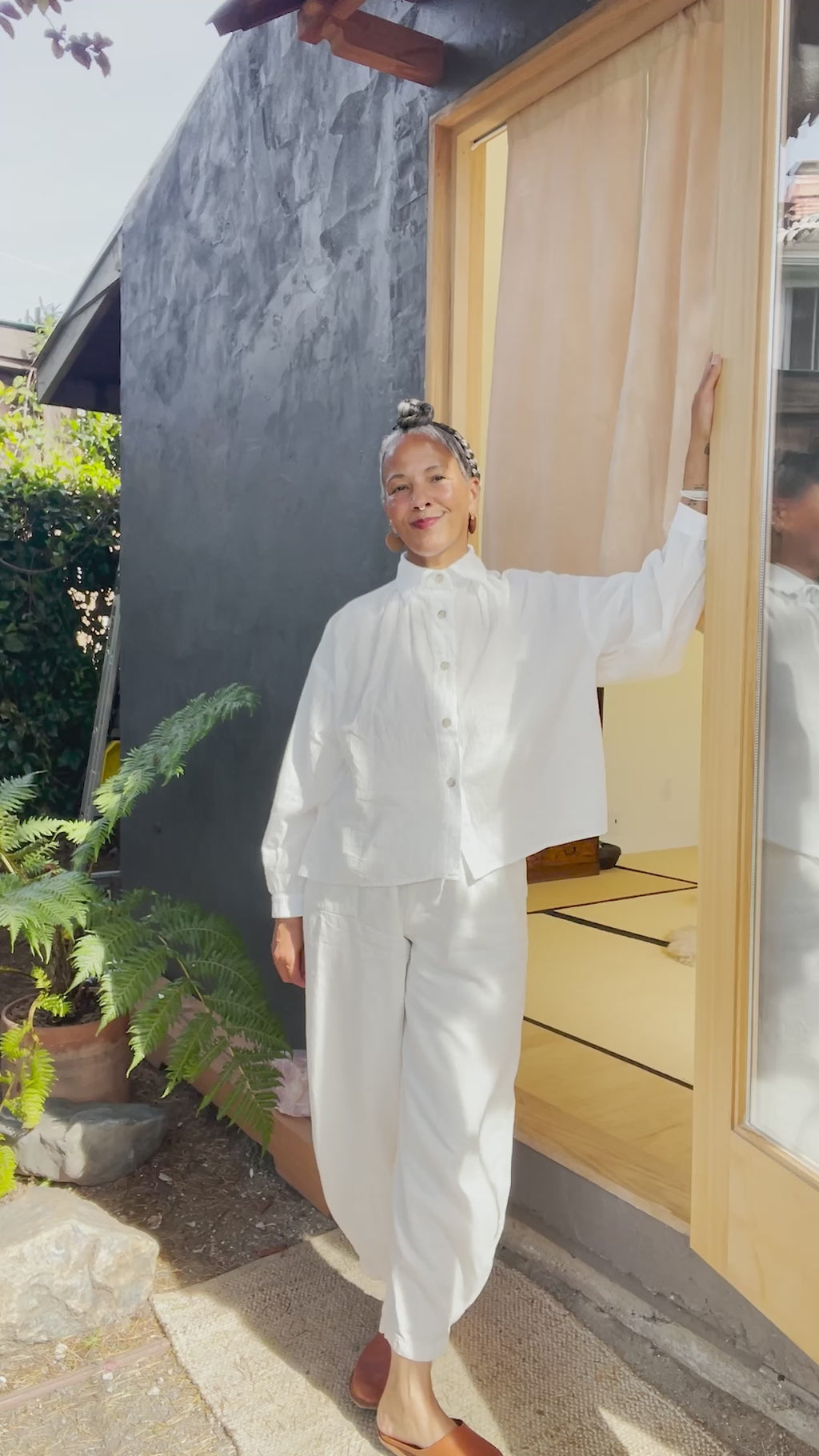 Video of model wearing Cropped Poet Blouse in White and Cadence Pants in White, showing different angles and closeups of shirt buttons, in front of grey building 