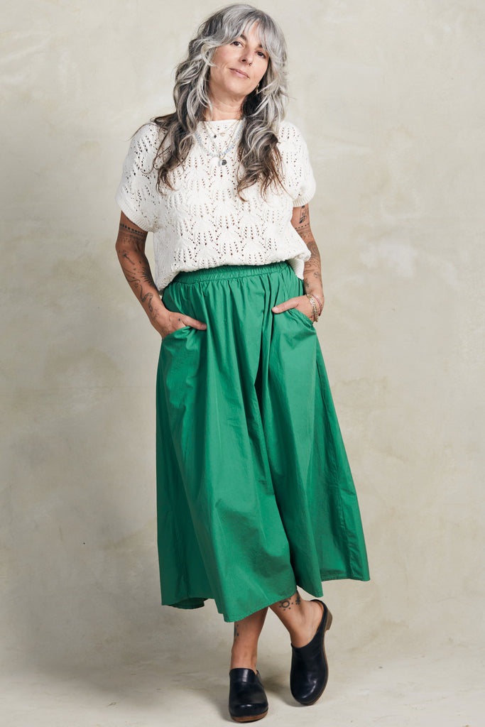 Front view of model wearing Rosey Sweater in Biscuit and Fawn Poplin Skirt in Fern, standing with hands in skirt pockets in front of white background