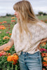 Model wearing Rosey Sweater in Biscuit and light wash jeans amongst foliage with orange flowers 
