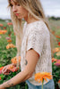 Model wearing Rosey Sweater in Biscuit and light wash jeans amongst foliage with orange flowers 