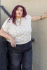 Model wearing Rosey Sweater in Biscuit and dark wash jeans standing in front of black and gray building