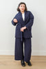 Front view of model wearing Sadie Coat in Indigo, Ayana Top in White, and Dover Pant in Indigo, tying belt for coat around waist in front of white background