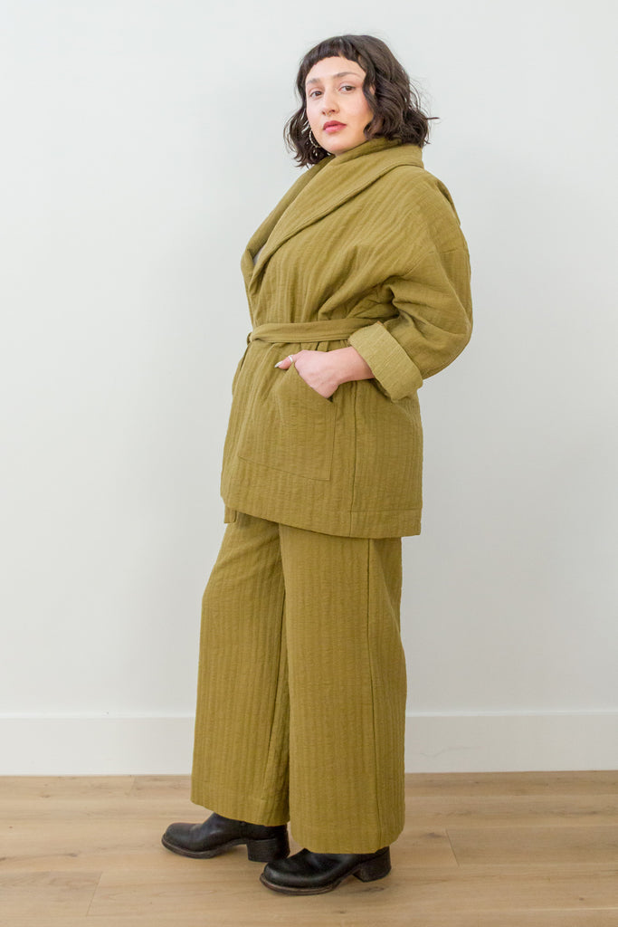 Side view of model wearing Sadie Coat in Fennel with the belt around the waist, Ayana Top in White, and Dover Pant in Fennel, with one hand in jacket pocket in front of white background