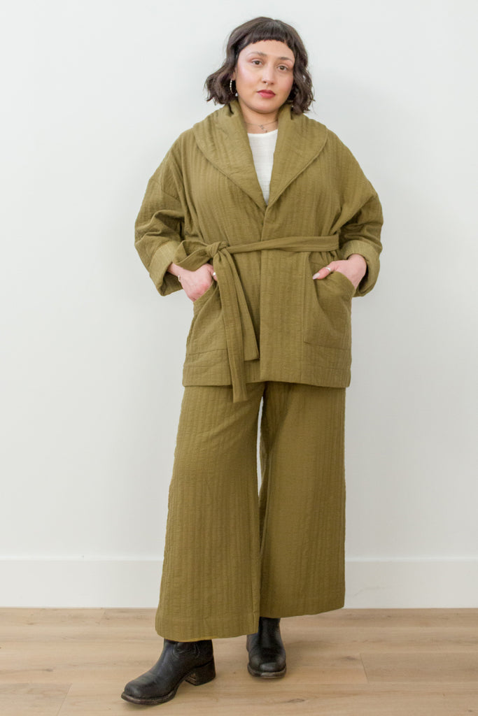 Front view of model wearing Sadie Coat in Fennel with the belt around the waist, Ayana Top in White, and Dover Pant in Fennel, with hands in jacket pockets in front of white background