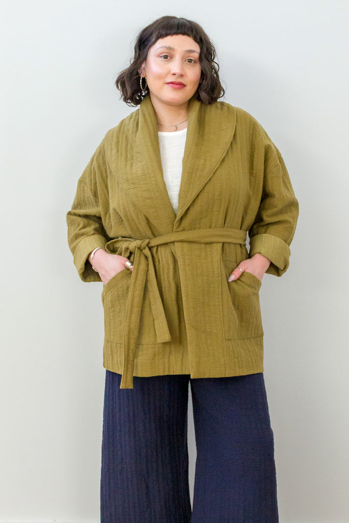 Front view of model wearing Sadie Coat in Fennel with the belt around the waist, Ayana Top in White, and Dover Pant in Indigo, with hands in jacket pockets in front of white background