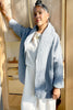 Woman wearing a stone blue Sadie Coat - textured jacket over a white top and pants, standing outdoors.