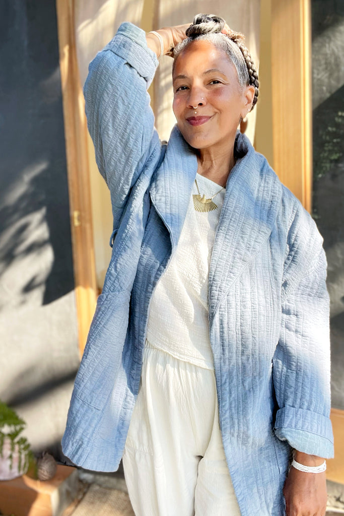 Woman wearing a stone blue Sadie Coat - textured jacket over a white top and pants, standing outdoors.