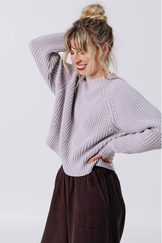Side view of model wearing Sonnie Sweater in Mist and Atlas Pant in Chocolate with one hand raised above head and one hand on hip in front of white background
