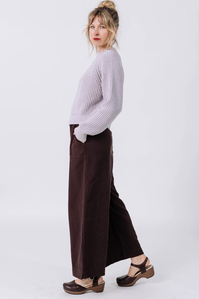 Side view of model wearing Sonnie Sweater in Mist and Atlas Pant in Chocolate, standing  in front of white wall