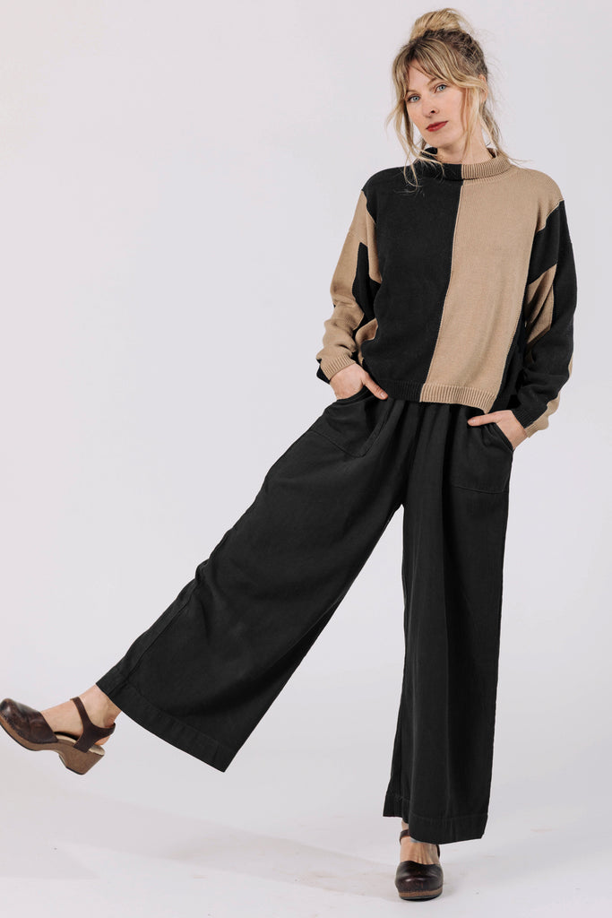 Front view of Atlas Pant in Black on model, paired with Sybil Sweater in Black/Tan, kicking a leg up in front of white background