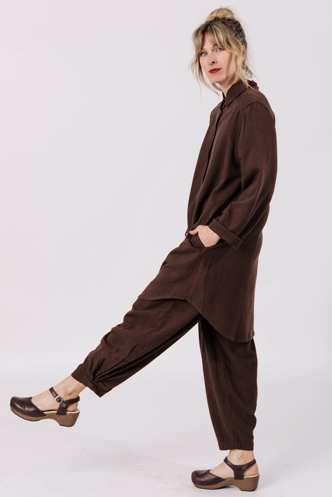 Side view of model wearing Workshirt and Cadence pants (both in Chocolate) standing with one leg kicking out in front of them, in front of white background
