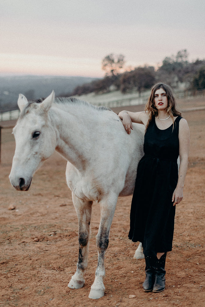 Standing model in tie waist sleeveless black skirt and black boots next to white horse