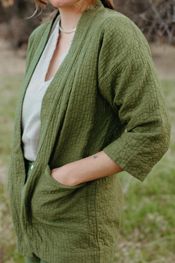 Close up photo of standing model with hands in pockets of green quilted jacket paired with white v-neck top and green pants