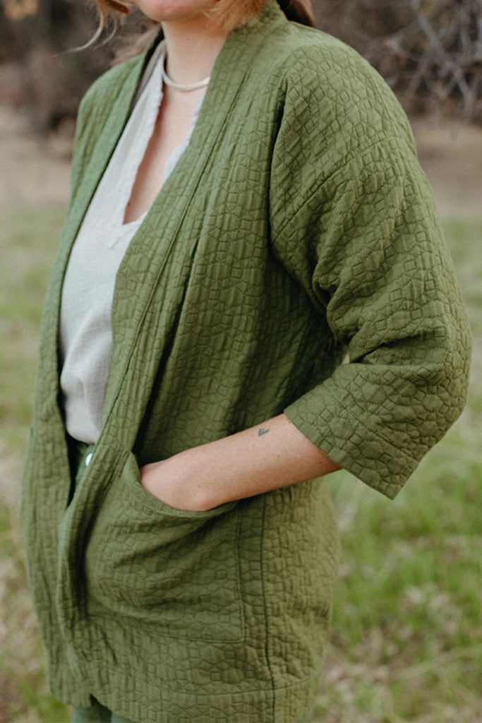 Close up photo of standing model with hands in pockets of green quilted jacket paired with white v-neck top and green pants