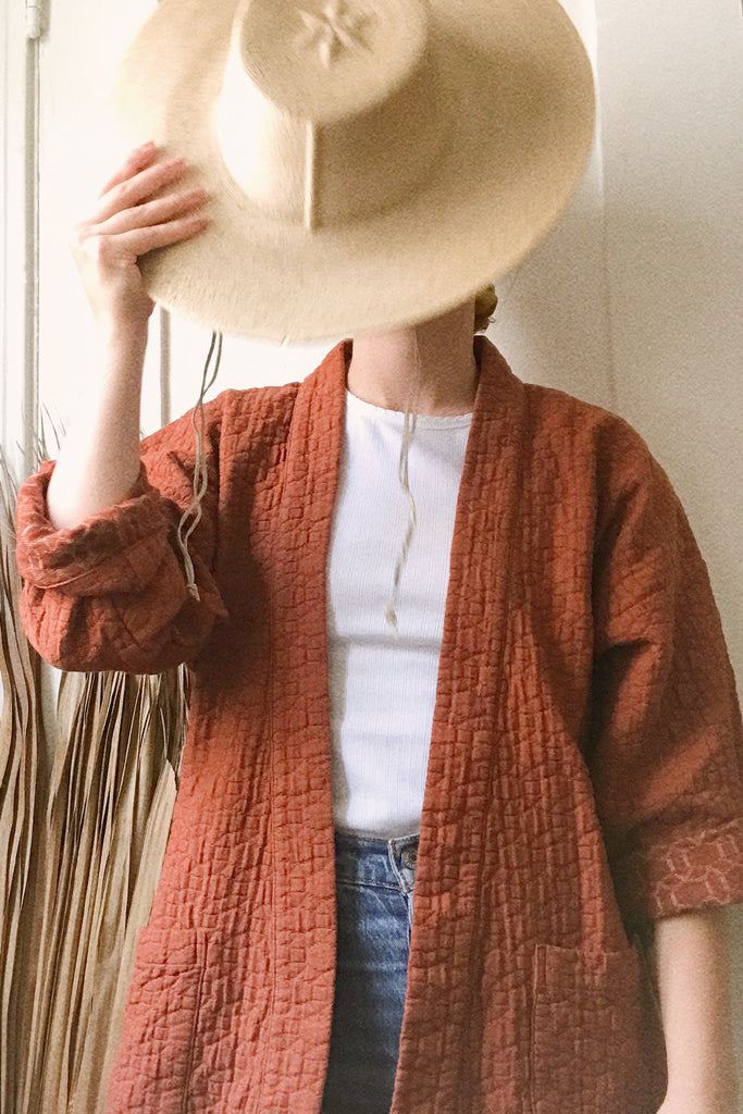 model in quilted rust colored jacket, paired with white top, jeans, and straw hat