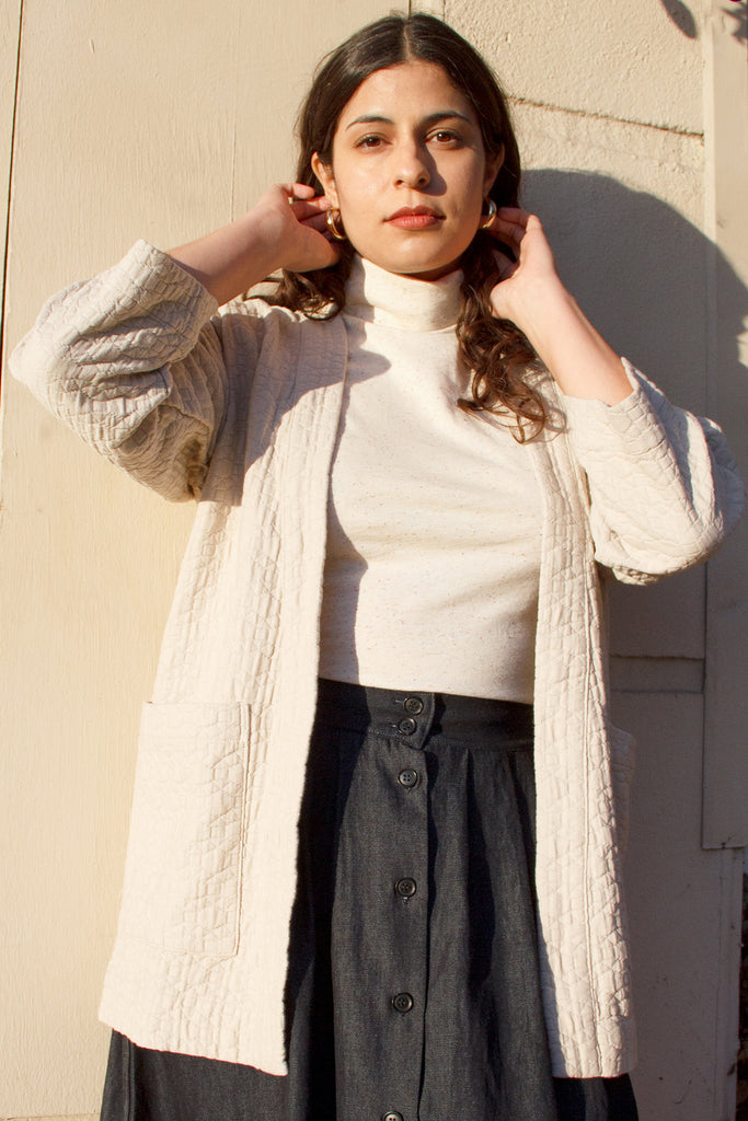 Beige quilted jacket on standing model, paired with white turtleneck top and dark denim button up maxi skirt