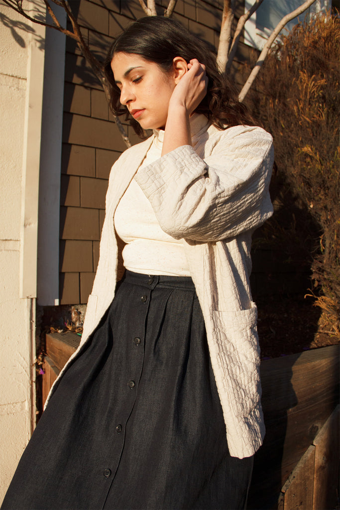 Side facing model with beige quilted jacket, paired with white top and dark denim button front maxi skirt